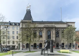 Das Berliner Ensemble von außen an einem Sommertag. Im Vordergrund der Bertolt-Brecht-Platz mit Statue von Bertolt Brecht, auf dem Dach des Theatergebäudes am Schiffbauerdamm ist der Berliner Ensemble-Kreisel zu sehen.