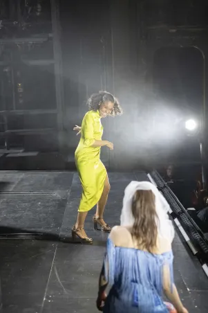 Cynthia Micas in einem gelben Kleid auf der Bühne, im Vordergrund von hinten zu sehen: Laura Balzer