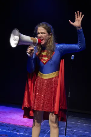 Frau in Superheldenkostüm auf dunkler Bühne, hebt eine Hand und spricht wütend in ein Megaphon