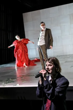 Szene mit drei Personen auf einer Bühne aus Holz. Eine Person vorne mit Mikrofon und dunklem Anzug, im Hintergrund eine Person in rotem Kleid und eine in Anzug mit Sonnenbrille.