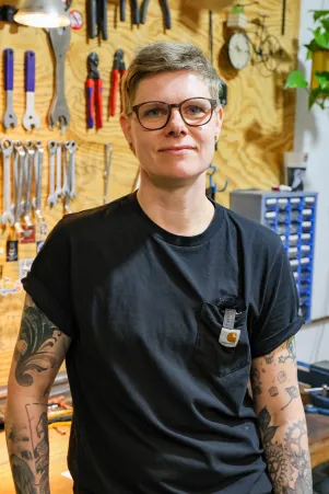 Werkstattleiterin Jenny Andersen an ihrem Arbeitsplatz. Sie trägt ein schwarzes Shirt, eine Brille, ihre Armtattoos sind zu erkennen. 