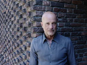 Schauspieler Christian Berkel in blaugrauem Hemd vor einer Backsteinmauer, schaut in die Kamera. 