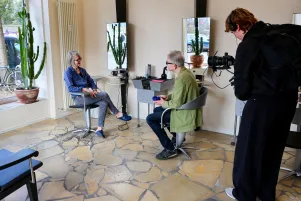Backstagefoto vom Dreh: Es ist ein Friseursalon von innen abgebildet, Oliver Reese und Sabine Lepenis sitzen einander  auf Stühlen gegenüber. Rechts ist der Kameramann zu erkennen. 