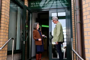 Jacqueline Dey und Oliver Reese stehen vor der offenen Eingangstür des Bestattungsinstituts Berg, sie begrüßen sich. 