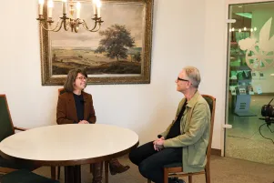Jacqueline Dey und Oliver Reese an einem runden Tisch im Büro der Bestattungsberaterin. Im Hintergrund ist ein Bild einer Landschaft zu erkennen, von der Decke hängt ein Kronleuchter. 