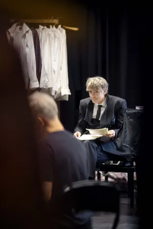 Jens Harzer im Probenkostüm mit Textbuch in der Hand, im Hintergrund Kleiderbügel, an denen weiße Hemden hängen.