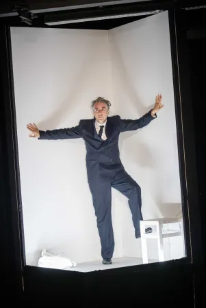Jens Harzer im Bühnenbild. Er befindet sich in einem schmalen hohen und weißen Kasten, er trägt einen dunklen Anzug. 
