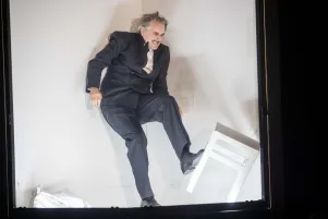 Jens Harzer im Probebühnenbild, er ist in Bewegung und tritt mit dem linken Fuß einen weißen Hocker weg. 