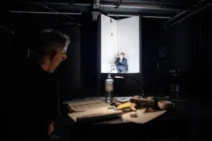 Jens Harzer kauert im weiß beleuchteten Bühnenbild, im vorderen, dunkleren, Bildteil ist Oliver Reese beobachtend im Profil zu erkennen. 