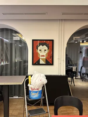 Die Platzierung des Portraits von Helene Weigel zwischen zwei Rundbögen in der Kantine. Im vorderen Bildbereich steht eine Trittleiter, darauf ein Eimer mit Arbeitsmaterialien.