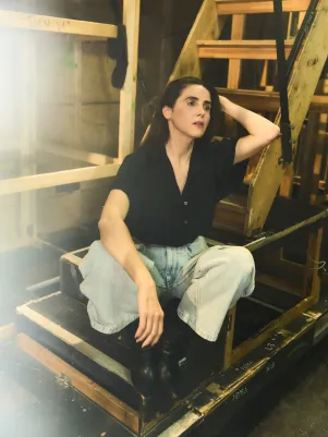 Schauspielerin Marina Galic sitzt in heller weiter Jeans und schwarzer Bluse auf einer Treppe unter der Bühne, fährt sich mit der Hand durchs Haar.