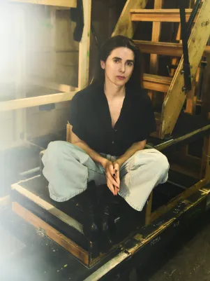 Schauspielerin Marina Galic sitzt in heller weiter Jeans und schwarzer Bluse auf einer Treppe unter der Bühne, schaut direkt in die Kamera.