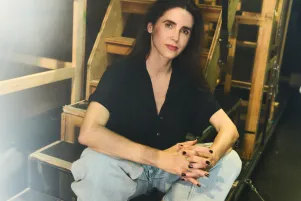 Schauspielerin Marina Galic sitzt in heller weiter Jeans und schwarzer Bluse auf einer Treppe unter der Bühne, schaut direkt in die Kamera.