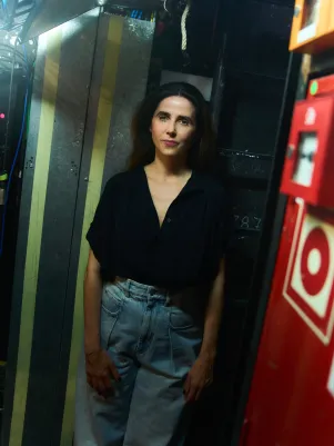 Schauspielerin Marina Galic steht in heller weiter Jeans und schwarzer Bluse hinter der Bühne, schaut direkt in die Kamera. Im Vordergrund ist an der Seite ein Feuer-/Brandmelder zu sehen.