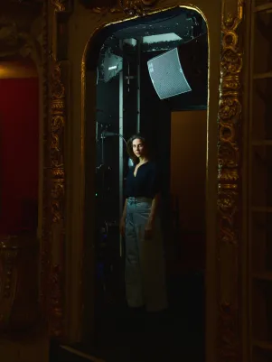 Schauspielerin Marina Galic steht in heller Jeans und schwarzer Bluse in einer Loge im Zuschauerraum des Großes Hauses. Sie schaut im Halbprofil in die Kamera, die Verzierungen des Raumes bilden einen Rahmen um sie.