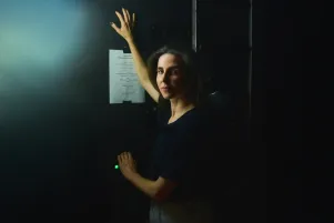 Schauspielerin Marina Galic in heller Jeans und schwarzer Bluse steht im Halbprofil vor schwarzer Wand. Sie lehnt mit den Händen gegen die Wand, darauf ist ein weißes DIN A4 Blatt zu erkennen. 