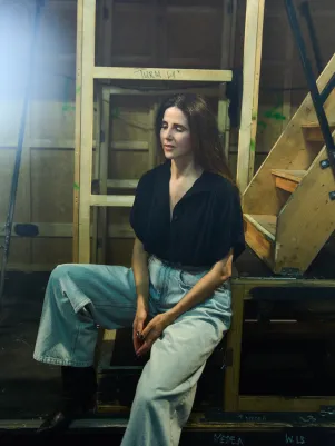 Schauspielerin Marina Galic sitzt in heller weiter Jeans und schwarzer Bluse auf einer Leiter unter der Bühne.