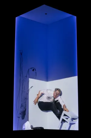 Jens Harzer in weißem Hemd, Krawatte und schwarzer Anzughose. In einem blau beleuchteten Würfel lehnt er sich an die Wand, mit den Füßen auf einem weißen Hocker. Sein Gesicht ist beschmiert, die Wand des Würfels bekritzelt.