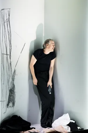 Schauspieler Jens Harzer steht leicht gebeugt in schwarz gekleidet an eine Wand gelehnt, hält eine Flasche in der Hand. Im Hintergrund sind skizzenhafte Linien an der Wand, auf dem Boden um ihm liegen Klamotten verteilt.