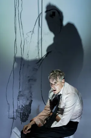 Schauspieler Jens Harzer sitzt gebeugt in weißem Hemd und schwarzer Krawatte auf einem weißen Hocker. Er hält ein Mikrofon. Die Wand hinter ihm ist bekritzelt und fängt seinen Schatten ein. Sein rechter Ärmel ist blutverschmiert.