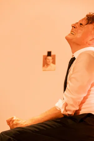 Schauspieler Jens Harzer sitzt in orangenem Licht, er weint und hat eine angespannte Körperhaltung. Unscharf im Hintergrund ist ein Foto an der Wand zu erkennen.