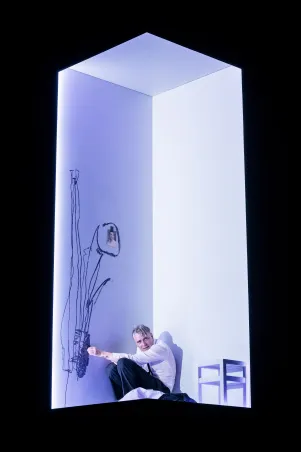 Schauspieler Jens Harzer kauert in einem beleuchteten Würfel auf dem Boden, neben ihm ist ein weißer Hocker, auf dem Boden liegen Klamotten, er skizziert an der Wand, daran klebt eine Fotografie.