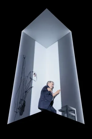 Fotografiert von unten: Schauspieler Jens Harzer lehnt an die Wand in hell beleuchteten Würfel in dunkler Kleidung. Mit der Hand fährt er die Wand entlang. An der Wand hinter ihm ist eine grobe Skizze, daneben klebt eine Fotografie, an der Kante des Würfels steht ein weißer Hocker.