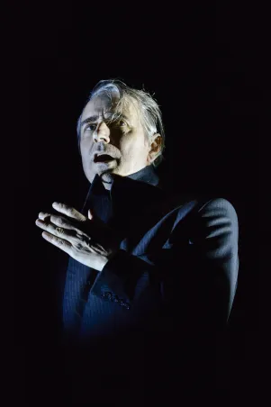 Jens Harzer in dunklem Sakko, spricht, hält eine Zigarette in der Hand. Die Bühne ist dunkel, lediglich Harzer ist beleuchtet.