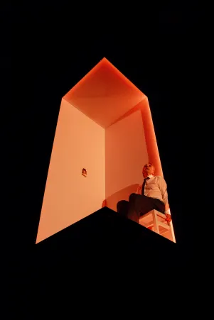 Schauspieler Jens Harzer sitzt auf einem weißen Hocker am Rand eines orange beleuchteten Würfel auf der Bühne. Er trägt ein Hemd und eine Krawatte. An der Innenwand des Würfels klebt eine Fotografie, das Foto ist von unten aufgenommen.