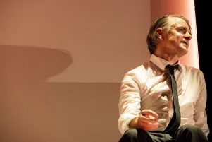 Schauspieler Jens Harzer sitzt in einem weißen Hemd und schwarzer Krawatte auf einer Bühne. Die Wand hinter ihm ist orange-rot beleuchtet, er spricht und schaut Richtung Zuschauerraum.