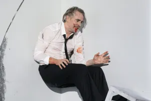 Schauspieler Jens Harzer lehnt mit dem Rücken gegen eine Wand und den Füßen auf einem weißen Hocker, er trägt ein weißes Hemd, eine schwarze Krawatte und eine schwarze Hose. Seine Hände und Gesicht sind mit Blut verschmiert. Die Wand hinter ihm ist bekritzelt.