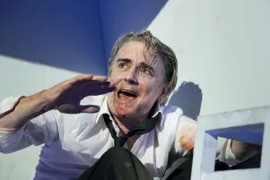 Schauspieler Jens Harzer sitzt auf dem Boden, trägt ein weißes Hemd und eine schwarze Krawatte, mit blutigen Flecken auf der Kleidung und im Gesicht. Er hat eine Hand erhoben und spricht. Im Vordergrund ist unscharf ein weißer Hocker zu erkennen.