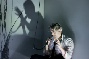 Schauspieler Jens Harzer sitzt in weißem Hemd und schwarzer Krawatte an eine Wand gelehnt. Er spricht in ein Mikrofon. Die Wand neben ihm ist bekritzelt und fängt seinen Schatten ein.