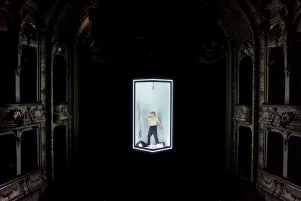 Schauspieler Jens Harzer steht in einem beleuchteten rechteckigen Rahmen auf einer Bühne. Der Hintergrund ist dunkel, die Bühne hat barocke Verzierungen. Er ist oberkörperfrei, trinkt aus einer Flasche, auf dem Boden um ihm liegen Klamotten und die Wand hiner ihm ist bekritzelt. An der Decke des Würfels über ihm hängt ein Mikrofon.