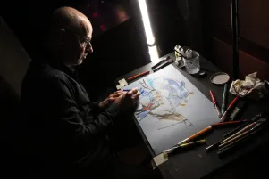 Der Künstler Reinhard Kleist ist seitlich fotografiert worden. Er befindet sich im Halbdunkel, das Papier vor ihm ist beleuchtet. Er malt mit bunten Farben.
