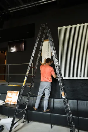 Ein mann steht auf einer Sitzgelegenheit und bringt eine Schablone an der Wand an, vor ihm steht eine hohe Trittleiter. 
