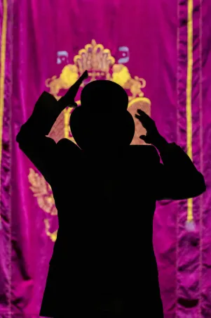 1)	Silhouette einer Person mit Hut und erhobenen Händen vor einem violetten Vorhang mit goldfarbenem Ornament. 