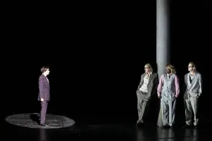 10)	Eine Person im karierten Anzug steht auf einem beleuchteten runden Bereich, drei weitere Personen lehnen an einer Säule, alle tragen helle Anzüge.