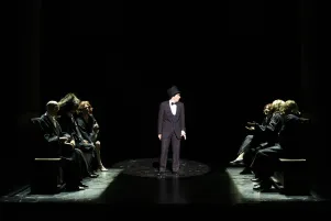 12)	Eine Person in dunklem Anzug und Zylinder steht im Scheinwerferlicht auf einer Bühne, umgeben von sechs sitzenden Personen in dunkler Kleidung auf Bänken.