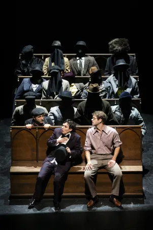 15)	Zwei Männer sitzen vorne auf einer Holzbank, dahinter mehrere Reihen mit Figuren in Anzügen und Hüten, deren Gesichter durch schwarze Schleier verdeckt sind.