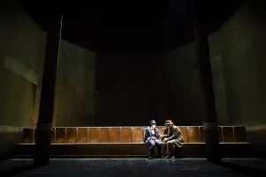 17)	Zwei Männer in Anzügen und Hüten sitzen auf einer langen Holzbank in einem dunklen, hohen Raum mit Säulen und großen Wandtafeln.