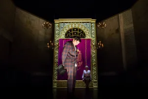 19)	Mann in kariertem Anzug und Hut steht vor einem goldverzierten Schrein mit rotem Samtvorhang, Frau sitzt auf dem Schreib, umgeben von Kronleuchtern.