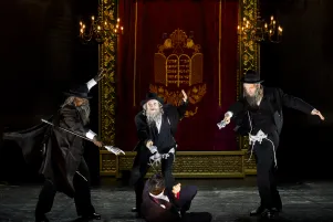 20)	Drei Männer in schwarzen Anzügen und Hüten mit weißen Schläfenlocken und langen Bärten umringe tanzend eine am Boden liegende Person. Im Hintergrund ein roter Vorhang mit goldener Verzierung.