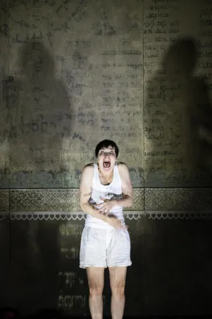 3)	Frau in weißem ärmellosem Hemd und weißen Shorts steht vor einer Wand mit alten, verblassten Schriftzeichen und Ornamenten.