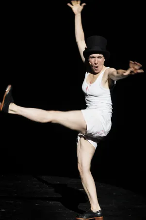 6)	Person in weißem ärmellosem Oberteil und Shorts, trägt schwarzen Zylinderhut und schwarze Schuhe, steht auf einem Bein und hebt das andere gestreckt nach vorne.