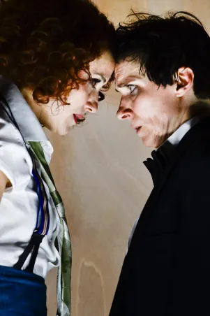 7)	Zwei Personen stehen sich Stirn an Stirn gegenüber, eine trägt ein weißes Hemd mit Hosenträgern und eine grün gemusterte Krawatte, die andere einen schwarzen Anzug mit weißem Hemd.