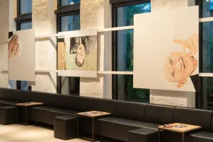 Es sind drei Leinwände zu erkennen, auf denen sich stilisierte Portraits befinden. Sie sind vor den Fenstern des Foyers im Neuen Haus befestigt. 