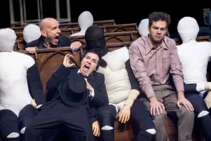 Kathrin Wehlisch, Paul Herwig und Gabriel Schneider sitzen zwischen Puppen auf harten Bänken, Kathrin Wehlisch und Paul Herwig schreien erschrocken. 