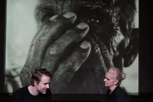 Inszenierungsfoto von Srebrenica, links im Bild Jens Harzer, rechts Vernesa Berbo, die zueinander gewandt sitzen. Im Hintergrund eine schwarz-weiß Projektion einer Nahaufnahme eines Mannes, der sich die Hände vors Gesicht hält