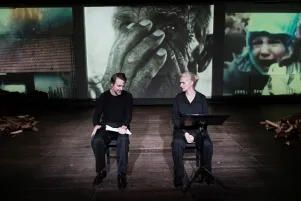 Inszenierungsfoto von Srebrenica, links im Bild Jens Harzer, rechts Vernesa Berbo, die zum Publikum gewandt auf der Bühne sitzen. Mittig im Hintergrund eine schwarz-weiß Projektion einer Nahaufnahme eines Mannes, der sich die Hände vors Gesicht hält, links der Ausschnitt einer Aufnahme eines brennenden Hauses, rechts ein weinendes Kind. Rechts und links auf der Bühne liegen kleine Haufen Holzscheite.